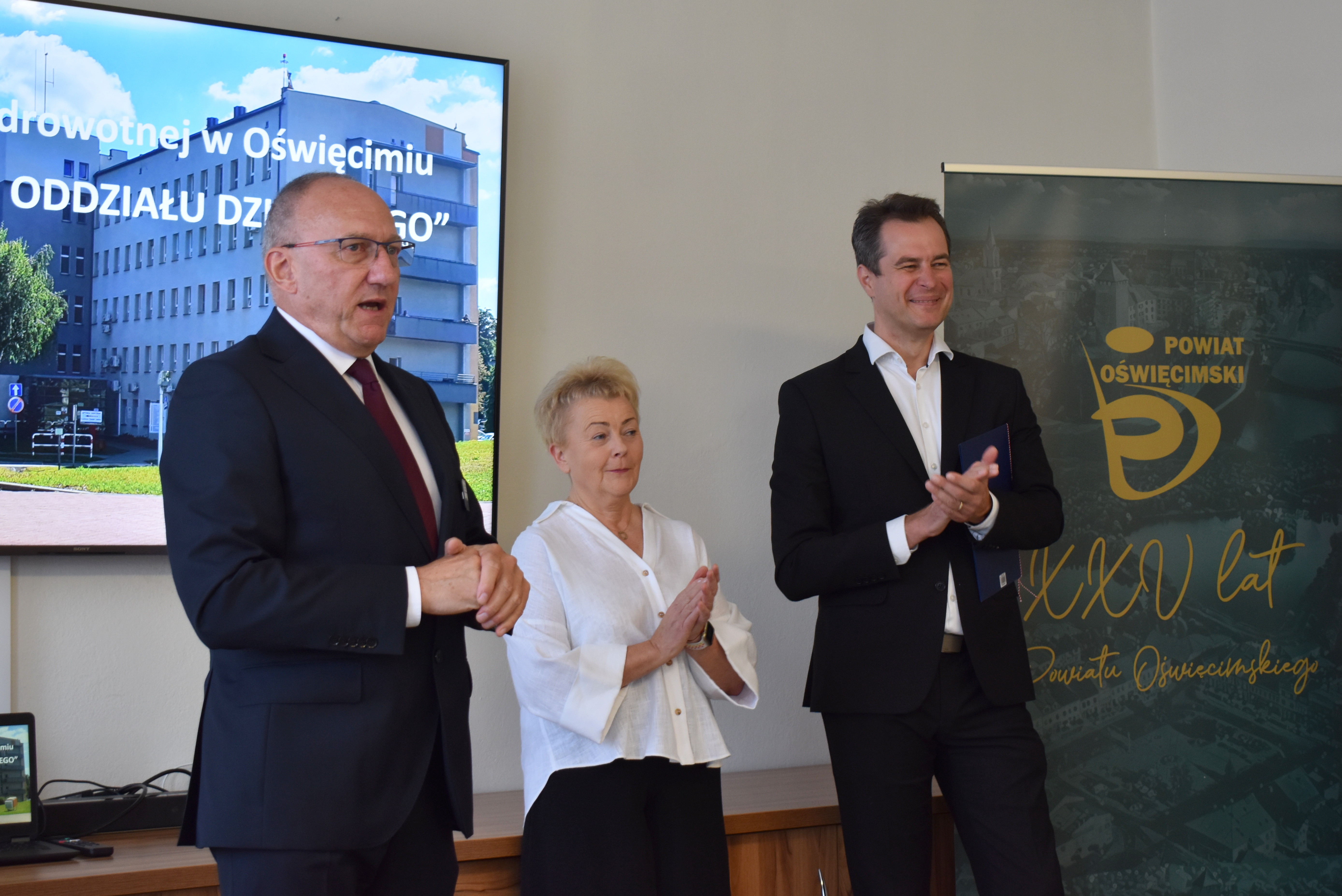 Dyrektor szpitala, ordynatorka oddziału dziecięcego i starosta oświęcimski stoją na tle ekranu, na którym wyświetlono zdjęcie budynku szpitala