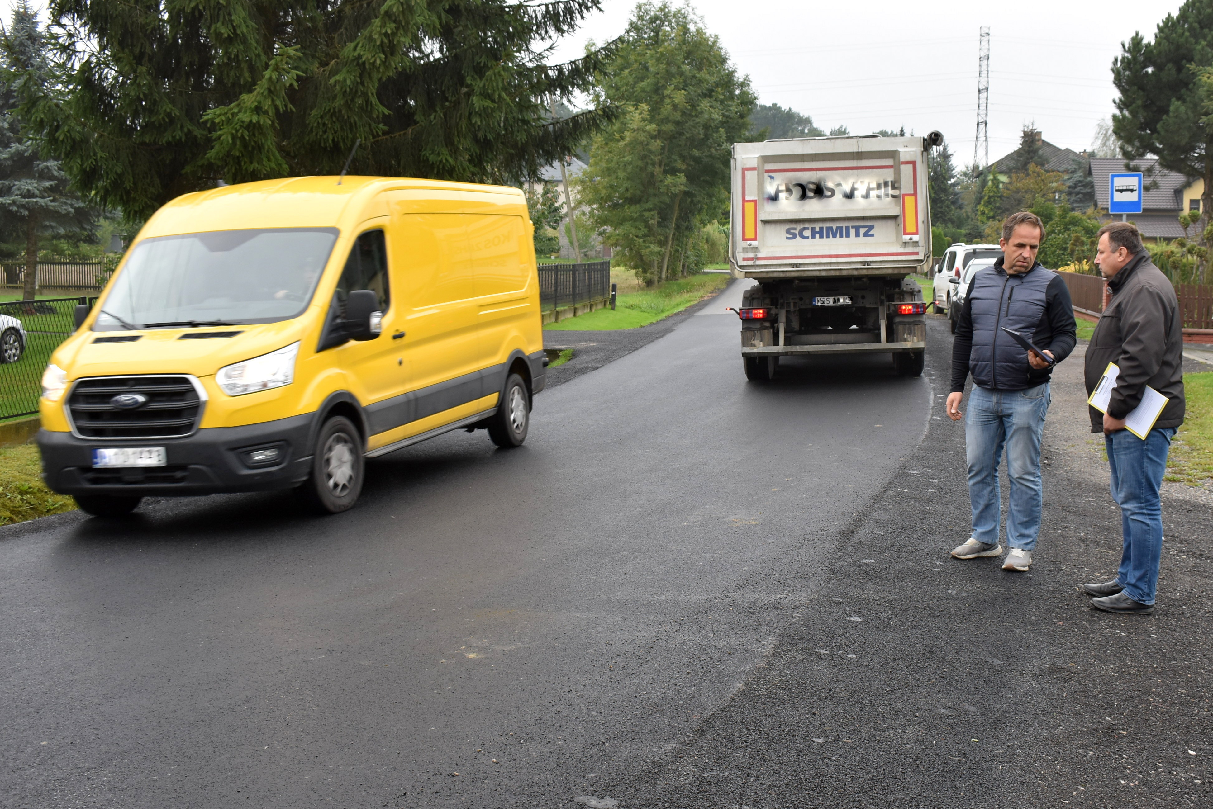 fragment wyremontowanej drogi, na której mijają się dwa pojazdy. Obok na poboczu stoją dwaj panowie